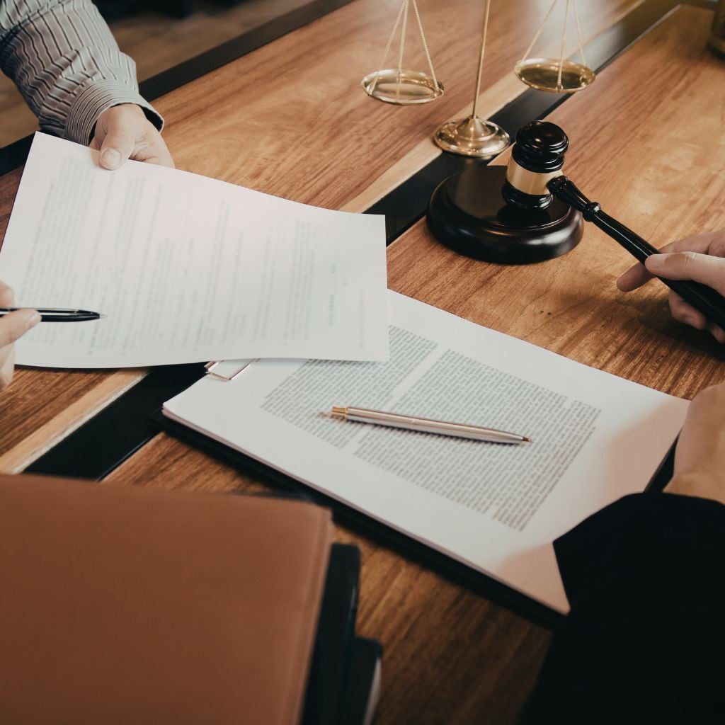 Two people review and sign documents at a wooden desk with a gavel, scales of justice, and legal paperwork present.