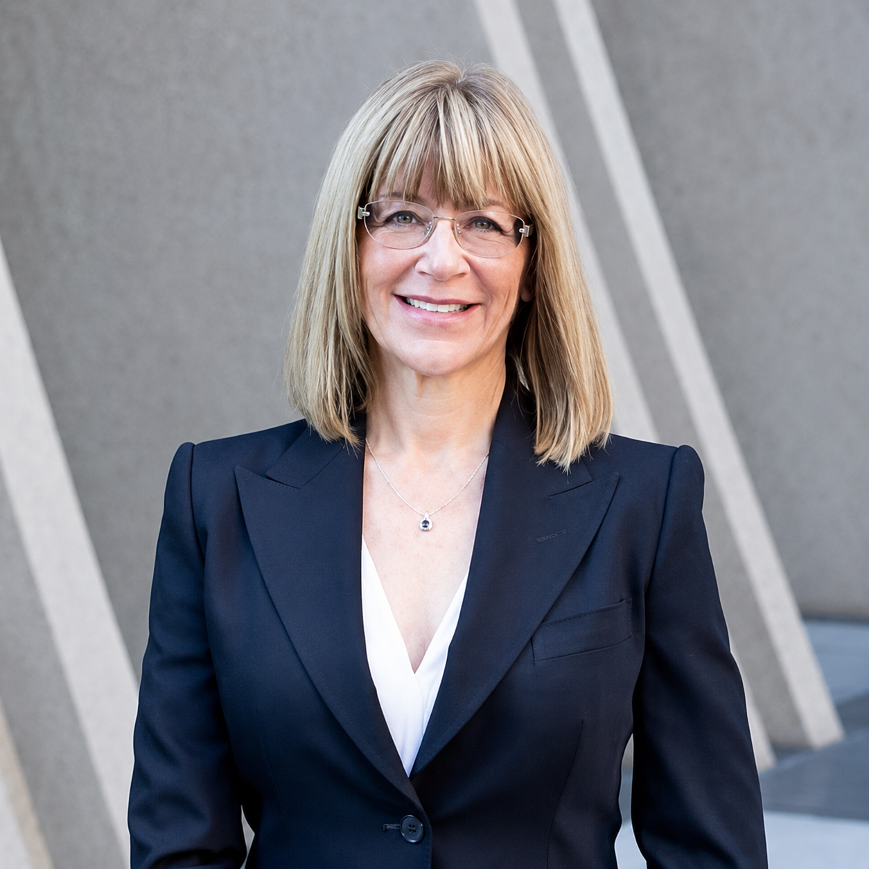 A woman with shoulder-length blonde hair, wearing glasses, a black blazer, and a white blouse, stands in front of a gray architectural background, smiling at the camera.
