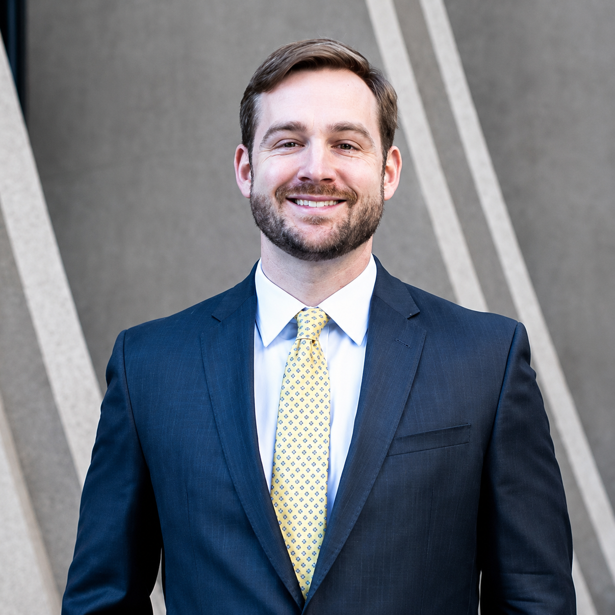 A man in a dark suit, light blue shirt, and patterned yellow tie stands outside in front of a gray wall with diagonal lines, smiling at the camera.