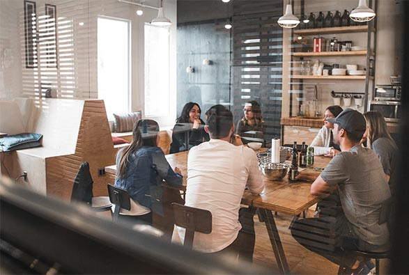 Seven people sit around a wooden table in a modern office meeting room, engaged in discussion, with shelves and windows visible in the background.
