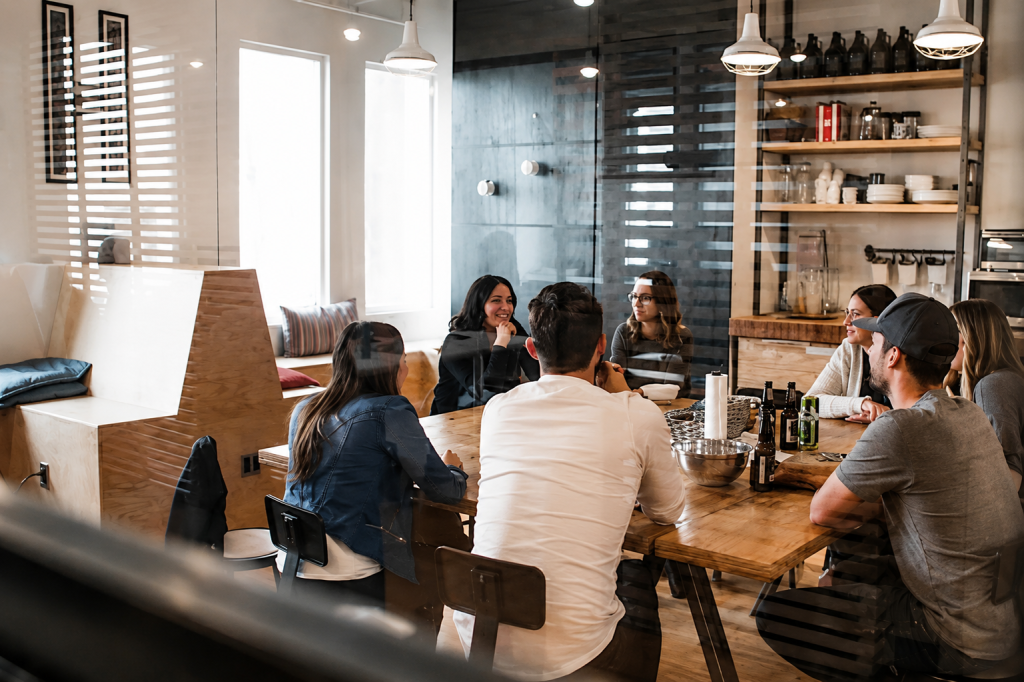 A group of people sit around a wooden table in a modern office meeting room, engaged in discussion. Shelves with dishes and bottles are visible in the background.
